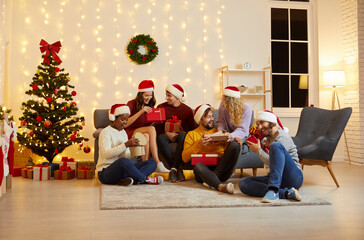Christmas mood. Group of young people exchange gifts and open gift boxes sitting by Christmas tree. Joyful and surprised multiracial people in Santa hats sit on sofa and on floor in cozy room at home.