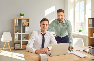Portrait of a two happy smiling confident business people wearing shirts looking cheerful at camera at the workplace sitting at the desk. Company employees and male coworkers working in office.