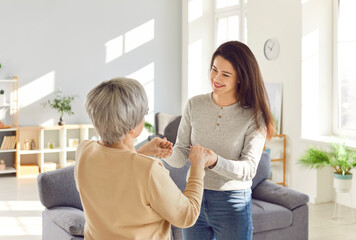 Portrait of a happy smiling adult daughter holding hands with her senior gray-haired old mother parent standing together in living room at home. Love, family and Mothers day concept.