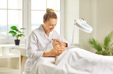 Young woman cosmetologist or dermatologist checking skin condition to a smiling girl client with special lamp in beauty spa salon. Facial massage, skin care treatment and cosmetology concept.