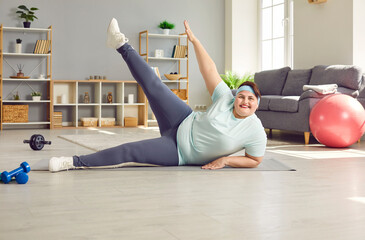 Happy overweight woman enjoying weight loss fitness workout at home. Beautiful smiling fat big large woman in sportswear lying on sports mat on floor doing gymnastics exercise with one leg raised up