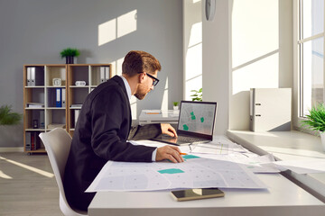 Professional cadastral surveyor in business suit sitting at table in modern office, using laptop computer, working with city structure design plan, looking at map, studying plot numbers and boundaries - 796959287