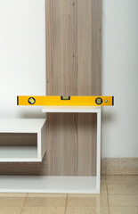Yellow spirit level on a wooden shelf that is being assembled. The shelf is against a white wall and the floor is beige. The level indicates that the horizontal piece of the shelf is perfectly aligned