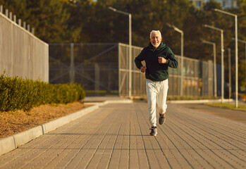 Senior runner man in a sporty ensemble enjoys a rejuvenating run in the city park at sunset. Smiling demeanor reflects the joy of physical activity, capturing active lifestyle through the jogging.