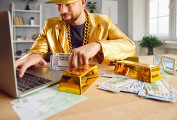 Funny happy Caucasian man in golden suit sitting at desk with money bills at home with laptop working or doing online shopping. Male person with cash and gold on table. Investment and success concept - 796966005