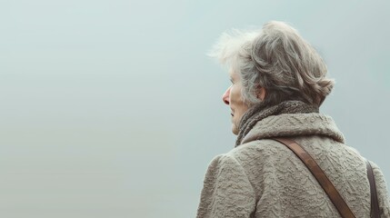 Thoughtful senior woman looking out at a foggy lake. She is wearing a warm winter coat and a scarf.
