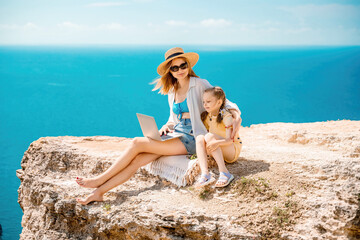 A woman and a child are sitting on a rock overlooking the ocean. The woman is using a laptop while the child looks on. Concept of relaxation and bonding between the two.