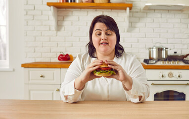 Overweight young woman eating hamburger while sitting at table in kitchen. Hungry plus size woman enjoying eating of delicious junk food. Unhealthy meal, overeating, extra calories - 796973000