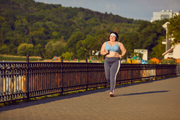 Happy smiling fat overweight chubby big large plump stout young woman in a sports bra and fitness pants having an outdoor jogging workout in summer and running on a bridge in a green sunny city park - 796975231