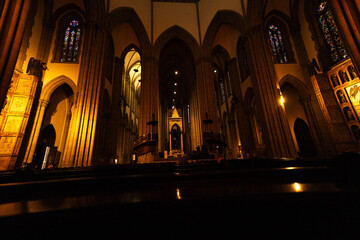 Interior de catedral com vitrais coloridos, bancos de madeira, colunas e arcos. 