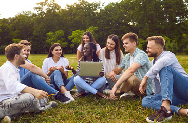 Group of happy diverse people friends sitting on the green grass in summer park with laptop, watching funny movie and talking with each other. Young guys and girls enjoying spending time together. - 796977673