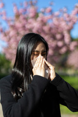 A young Asian woman sneezes in a springtime park, battling allergies amidst blooming flowers.