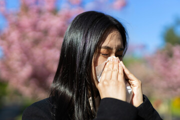 A young Asian woman, sick with allergies, sneezes in a park amidst springtime blossoms.