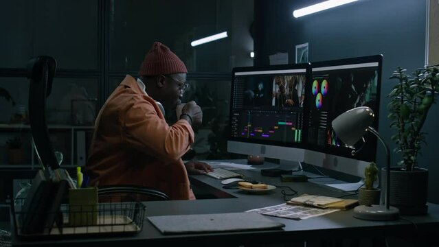 Medium full of black male video editor sitting at desk in dark postproduction studio, working on footage for zombie movie in specialist software on large computer screens, drinking coffee