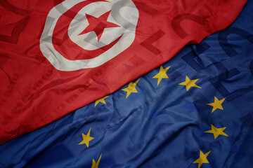 waving colorful flag of european union and flag of tunisia on a euro money banknotes background....