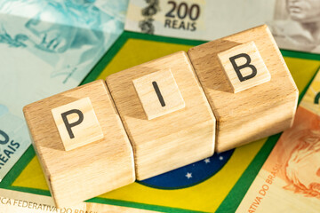 The word PIB (Gross Domestic Product) written on wooden cubes with some Brazilian real banknotes on yellow, green and blue national symbols of Brazil. Brazilian Portuguese language