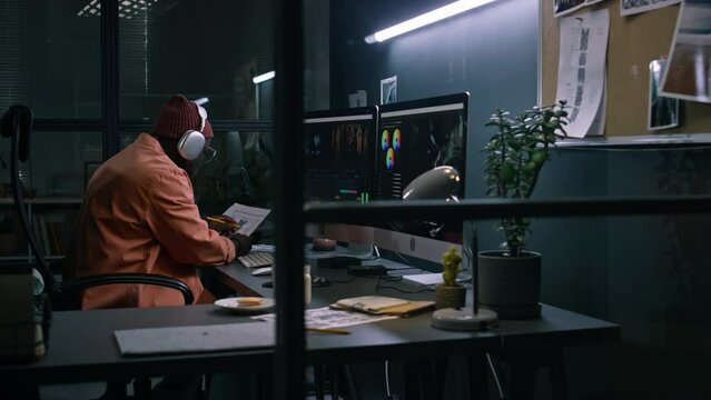 Medium full side of African American postproduction editor sitting at desk in office of filmmaking studio, working on raw footage in professional program on dual screen, then consulting movie script
