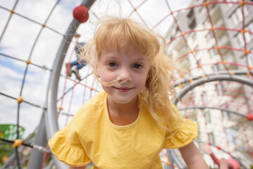 Cheerful child playing and having fun in an outdoor playground. A cute curly-haired smiling girl 6 years old in a yellow T-shirt climbs on a net. Active outdoor recreation for children in the city.