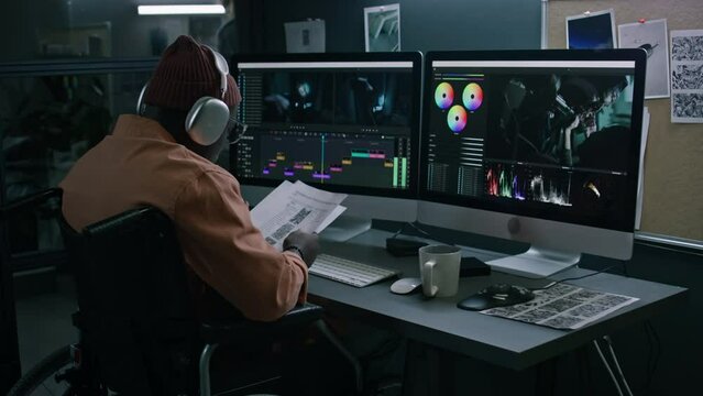 Medium full footage of professional African American male postproduction specialist with disability reading through film script, looking at frames, continuing to edit raw video material on computer