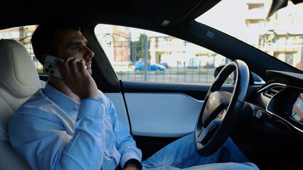 Male entrepreneur speaking by smartphone during riding on electrical auto with autopilot at urban road. Confident businessman talking on phone while riding an autonomous self driving electric car