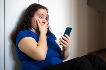 Cropped shot of a sad woman checking phone sitting on the floor in the living room at home with a dark background