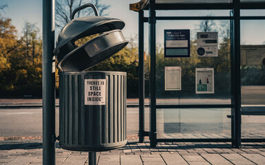 Public trash can, next to a bus stop with a request for recycling sticker 
