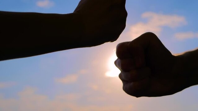 Close up view: man and woman hands silhouettes are bumping fists against the sunset or sunrise sky - sun lens flares. Informal greeting, giving dap, agreement, friendship and teamwork concept