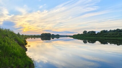 Fototapeta premium In the evening, the sun dips below the horizon and shines through the feathery clouds. The colorful sunset sky and trees are reflected in the calm river water. On the opposite bank is a forest