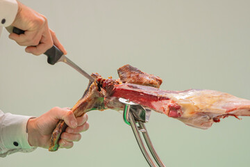 A person is cutting a piece of ham with a knife. The meat is cut into small pieces and is being held by a metal tool. Concept of preparation for cooking or eating the ham