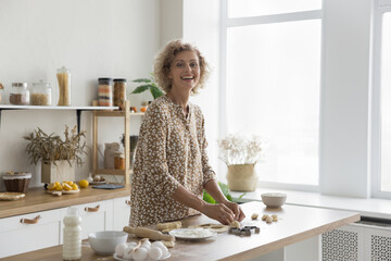 Laughing positive middle-aged housewife cooking in modern kitchen, staring at camera, standing at table with natural ecological products, kneading dough, shaping buns, make cookies on weekend at home