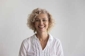 Pretty blond curly-haired mature woman posing in studio, having white-toothed smile advertises dental clinic services, head shot. Commercial offer for older people, medical insurance cover, midlife