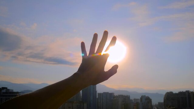 Silhouette of woman is waving her hand against the sunset, sunrise sky over the city, sun is shining through fingers - sun lens flares, close up. Freedom, summer and inspiration concept
