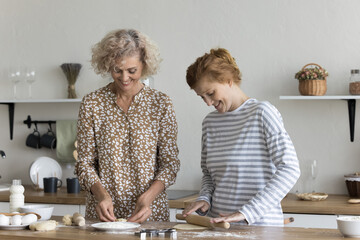 Smiling older and younger women cooking together in the kitchen. Happy mature female enjoy food preparation with young adult daughter, standing at table flattening homemade dough make cookies at home