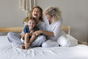 Two generations of Caucasian women, older granny and young mom share heart-warming moment, tickling little girl seated on bed, enjoy their friendly relations, boundless love and joy on cozy weekend