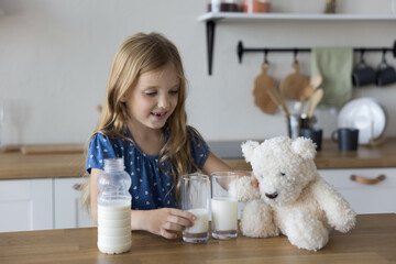 Pretty little girl poured two glasses of milk beverage for her and toy bear, caring for best friend health, play alone in kitchen. Calcium, vitamin D, and protein for bone growth and kid development