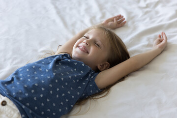 Above close up view pretty 5s Caucasian girl lying on cozy bed on white fresh bedsheets with stretched arms, smiling, enjoy carefree leisure, take daytime nap for good health and growth. Rest, comfort