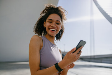 Young smiling woman in sportswear is resting after a workout outside while standing with smartphone