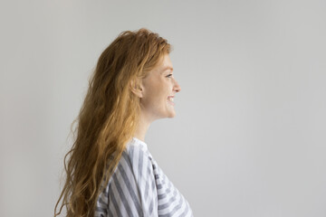 Side view of happy redhead pretty woman. Studio shot of single millennial female having long ginger hair and attractive appearance standing on gray studio wall background, staring straight and smiling