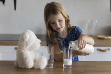 Little girl hold plastic bottle pours milk into two glasses for her and fluffy toy bear. Good life habit for children development and growth, vitamins and minerals necessary for kid overall health