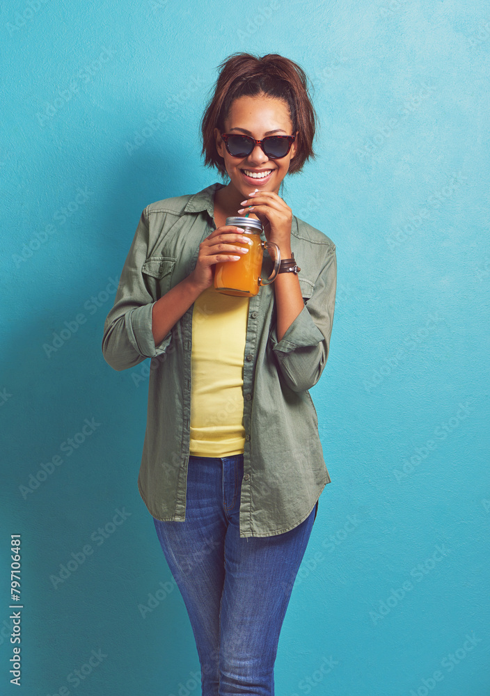 Poster Happy woman, portrait and sunglasses with orange juice for vitamin C or summer fashion on a blue studio background. Female person, brunette or model with smile for citrus drink or fresh beverage