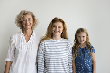 Three smiling beautiful relatives women posing on gray studio background, staring at camera, feel happy, showing family ties and unity. Heredity, from childhood to midlife, course of life, lifetime