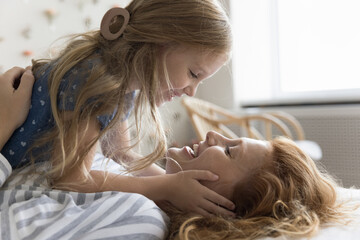 Close up joyful little daughter touch her loving mother face, lying together on bed, looking at...