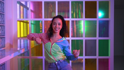 Happy hispanic girl in lively mood moving along music in neon light while wearing colorful cloth at night club. Street dancer making energetic footstep or movement while looking at camera. Regalement.