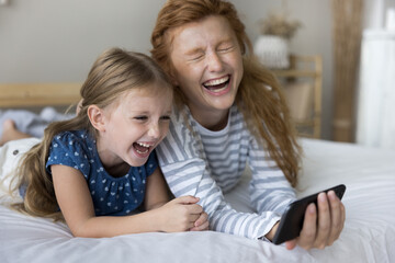 Joyful little cute girl relax in bed with mother on weekend at home, watch funny video or comedy movie, use smartphone online application, laughing. Happy family of two generation enjoy modern tech