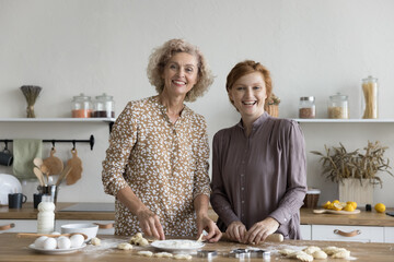 Intergenerational family, two women younger and older age cooking together in modern kitchen at home, standing at dining table, making dough, prepare cookies dessert for holiday, smile, look at camera