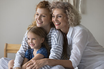 Happy middle-aged 60s woman hugging her young adult 30s daughter and cute little 5s granddaughter, smile, look away. Portrait of friendly loving family, good warm relationships, attachment and ties