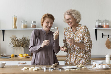 Intergenerational family, middle-aged mother and grown-up daughter cooking at home, young female swirls dough on finger, fooling, having fun while prepare homemade dessert for holiday in cozy kitchen