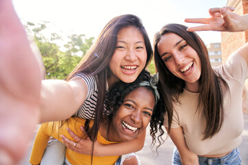 Group young multi-ethnic female taking smiling selfie portrait together outdoors. African woman giving piggyback ride to Asian girl on street. Excited nice friends posing for funny photo a sunny day