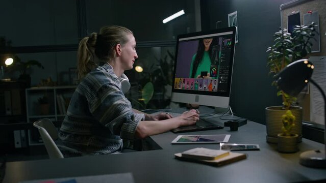 Medium side of blonde Caucasian female graphic designer working at computer desk in postproduction studio, editing advertising photos, retouching with stylus pen and digital tablet