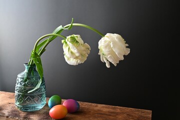 white tulips in a green vase standing on wood with dark background as well as colored eggs